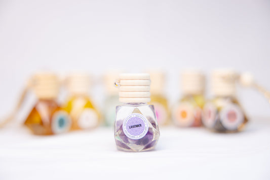 A small glass bottle of MounEssentials Untitled Aug8_06:37 with a wooden cap, holding purple essential oil, serves as an ideal car diffuser. In the background, various colorful bottles are blurred on a white surface.